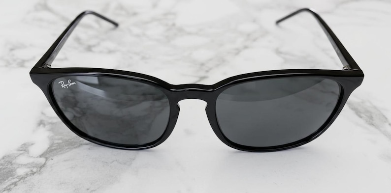Puede incluir: Gafas de sol negras con lentes grises oscuras. Las gafas tienen una montura redondeada y patillas finas. El logotipo de Ray-Ban es visible en la lente izquierda. Las gafas est&aacute;n sobre un fondo de m&aacute;rmol blanco y gris.