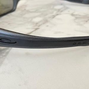 Może przedstawiać: Zbliżenie na parę ciemnoszarych okular&oacute;w przeciwsłonecznych z czarną oprawką. Logo Oakley jest widoczne na zauszniku. Soczewki mają odblaskową, iryzującą powłokę.