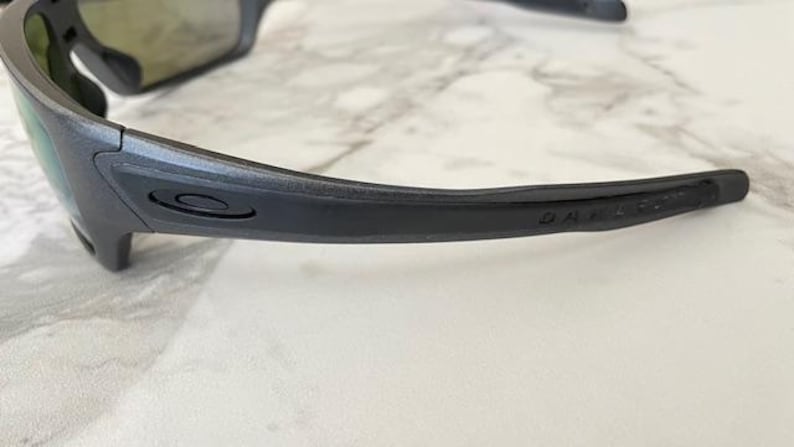 Może przedstawiać: Zbliżenie na parę ciemnoszarych okular&oacute;w przeciwsłonecznych z czarną oprawką. Logo Oakley jest widoczne na zauszniku. Soczewki mają odblaskową, iryzującą powłokę.