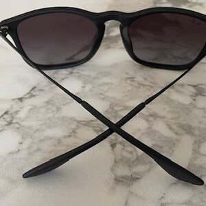 Puede incluir: Gafas de sol negras con lentes degradadas oscuras. Las monturas son negras y las patillas est&aacute;n cruzadas. Las gafas de sol descansan sobre una superficie de m&aacute;rmol blanco y gris.