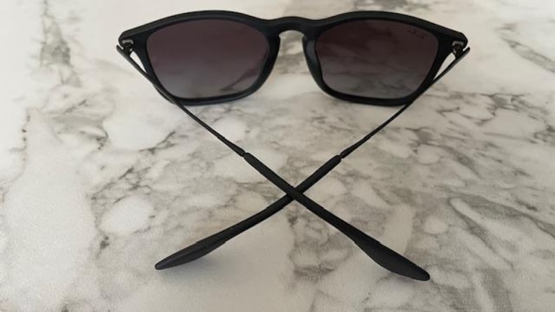 Puede incluir: Gafas de sol negras con lentes degradadas oscuras. Las monturas son negras y las patillas est&aacute;n cruzadas. Las gafas de sol descansan sobre una superficie de m&aacute;rmol blanco y gris.