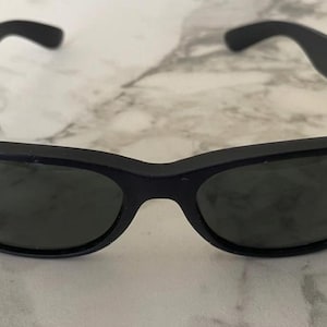 May include: Black sunglasses with dark lenses. The sunglasses have a classic, rectangular frame and are set against a marble background. The Ray-Ban logo is visible on the lens.