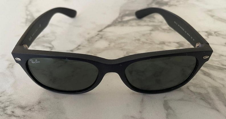 May include: Black sunglasses with dark lenses. The sunglasses have a classic, rectangular frame and are set against a marble background. The Ray-Ban logo is visible on the lens.