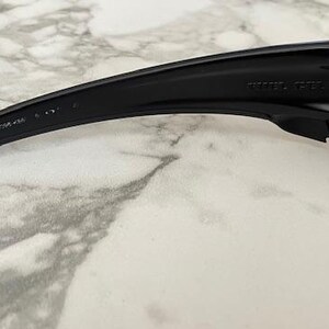 May include: Close-up of black sunglasses with a curved arm. The arm has text printed on it. The lens is a dark grey colour. The sunglasses are on a white and grey marble surface.