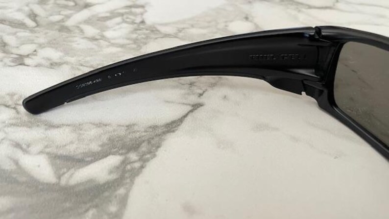 May include: Close-up of black sunglasses with a curved arm. The arm has text printed on it. The lens is a dark grey colour. The sunglasses are on a white and grey marble surface.