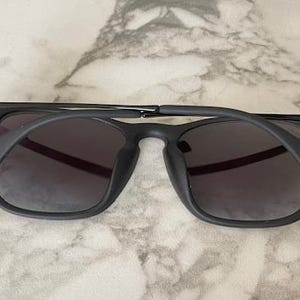 Puede incluir: Gafas de sol negras con lentes oscuras. Las gafas tienen una montura cuadrada y una fina barra met&aacute;lica en la parte superior. Las patillas son gruesas y se curvan hacia abajo para adaptarse a las orejas. Las gafas descansan sobre una superficie de m&aacute;rmol.