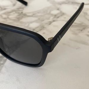 Puede incluir: Gafas de sol en negro mate con montura gruesa y lentes oscuras. Las patillas muestran dos finas rayas horizontales de color plateado cerca de la bisagra, y la montura tiene un acabado liso y sin reflejos con un puente distintivo.