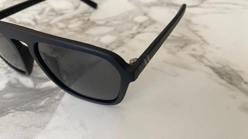 Puede incluir: Gafas de sol en negro mate con montura gruesa y lentes oscuras. Las patillas muestran dos finas rayas horizontales de color plateado cerca de la bisagra, y la montura tiene un acabado liso y sin reflejos con un puente distintivo.