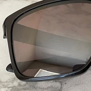 May include: Black sunglasses with a rectangular frame and tinted lenses. The sunglasses have a glossy finish and are set against a light-colored background. The lenses reflect the surrounding environment.