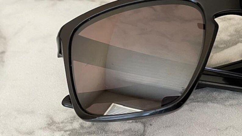 May include: Black sunglasses with a rectangular frame and tinted lenses. The sunglasses have a glossy finish and are set against a light-colored background. The lenses reflect the surrounding environment.