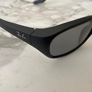 May include: Matte black sunglasses with dark grey lenses and a wrap-around frame design. The temples feature the white Ray-Ban logo printed on the side. A thin, flexible cord is attached to the end of the temple arms for secure placement.