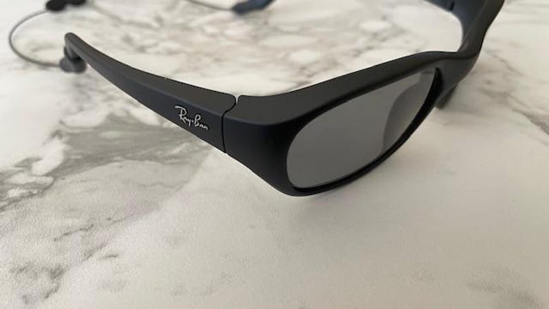 May include: Matte black sunglasses with dark grey lenses and a wrap-around frame design. The temples feature the white Ray-Ban logo printed on the side. A thin, flexible cord is attached to the end of the temple arms for secure placement.