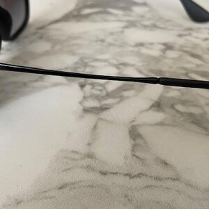 Puede incluir: Gafas de sol negras con lentes oscuras. Las gafas tienen un dise&ntilde;o elegante y moderno con una montura y patillas negras y finas. El logotipo de Ray-Ban es visible en la montura. Las gafas descansan sobre una superficie de m&aacute;rmol.