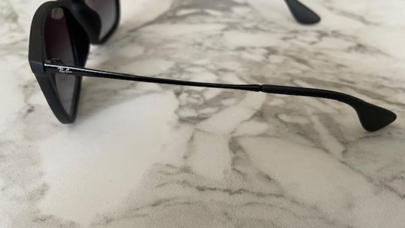 Puede incluir: Gafas de sol negras con lentes oscuras. Las gafas tienen un dise&ntilde;o elegante y moderno con una montura y patillas negras y finas. El logotipo de Ray-Ban es visible en la montura. Las gafas descansan sobre una superficie de m&aacute;rmol.