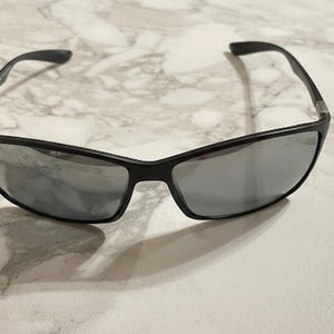 May include: Black sunglasses with gray lenses. The sunglasses have a rectangular shape and a sleek, modern design. The frames are a matte black, and the lenses are a reflective gray color. The sunglasses are resting on a white and gray marble surface.
