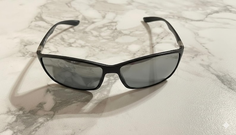 May include: Black sunglasses with gray lenses. The sunglasses have a rectangular shape and a sleek, modern design. The frames are a matte black, and the lenses are a reflective gray color. The sunglasses are resting on a white and gray marble surface.