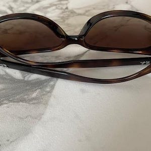 May include: Brown tortoiseshell sunglasses with dark lenses. The sunglasses have a classic, oversized frame design. The arms are curved and feature a subtle logo. The sunglasses are folded, resting on a light-colored surface.