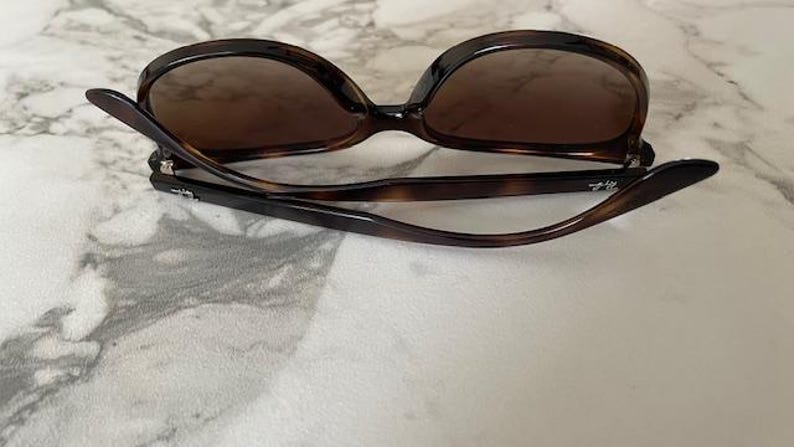 May include: Brown tortoiseshell sunglasses with dark lenses. The sunglasses have a classic, oversized frame design. The arms are curved and feature a subtle logo. The sunglasses are folded, resting on a light-colored surface.