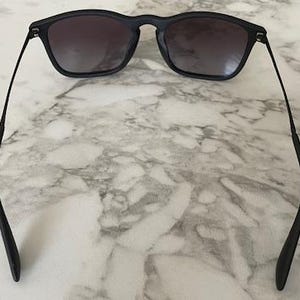 Puede incluir: Gafas de sol negras con lentes oscuras. Las gafas tienen una montura rectangular y patillas finas y curvadas. Las lentes son degradadas, pasando de oscuro en la parte superior a m&aacute;s claro en la inferior. Las gafas est&aacute;n sobre un fondo de m&aacute;rmol.