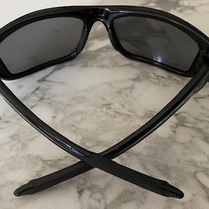 May include: Black sunglasses with dark gray lenses. The sunglasses have a sleek, modern design with a glossy finish. The arms of the sunglasses are crossed, showcasing their shape and style. The lenses appear to be polarized.