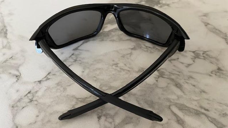 May include: Black sunglasses with dark gray lenses. The sunglasses have a sleek, modern design with a glossy finish. The arms of the sunglasses are crossed, showcasing their shape and style. The lenses appear to be polarized.