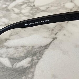 May include: Close-up of black sunglasses with a rectangular frame. The temple arm features the brand name and model information: 009102-6656 57 0 18 137 3N. The sunglasses are set against a marble-like surface.