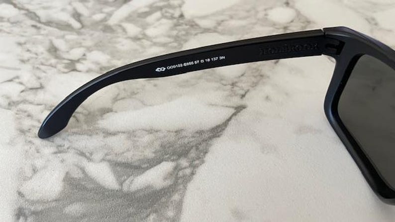 May include: Close-up of black sunglasses with a rectangular frame. The temple arm features the brand name and model information: 009102-6656 57 0 18 137 3N. The sunglasses are set against a marble-like surface.