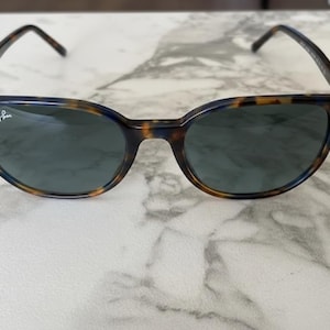 May include: Tortoiseshell-patterned sunglasses with dark lenses. The frames have a classic, rounded shape with a keyhole bridge. The Ray-Ban logo is visible on the left lens. The sunglasses are set against a marble background.