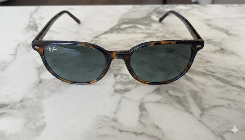May include: Tortoiseshell-patterned sunglasses with dark lenses. The frames have a classic, rounded shape with a keyhole bridge. The Ray-Ban logo is visible on the left lens. The sunglasses are set against a marble background.