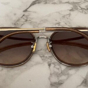May include: Brown sunglasses with a gold-colored bridge and arms. The lenses are a gradient brown, and the frame is a light brown. The arms have a glittery finish. The sunglasses are set against a marble background.