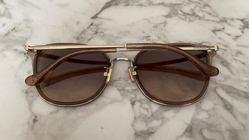 May include: Brown sunglasses with a gold-colored bridge and arms. The lenses are a gradient brown, and the frame is a light brown. The arms have a glittery finish. The sunglasses are set against a marble background.