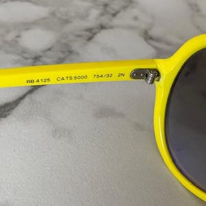 May include: Close-up of bright yellow sunglasses with dark lenses. The arm of the glasses displays the text "RB 4125 CATS 5000 754/32 2N". The sunglasses have a classic, rounded shape.