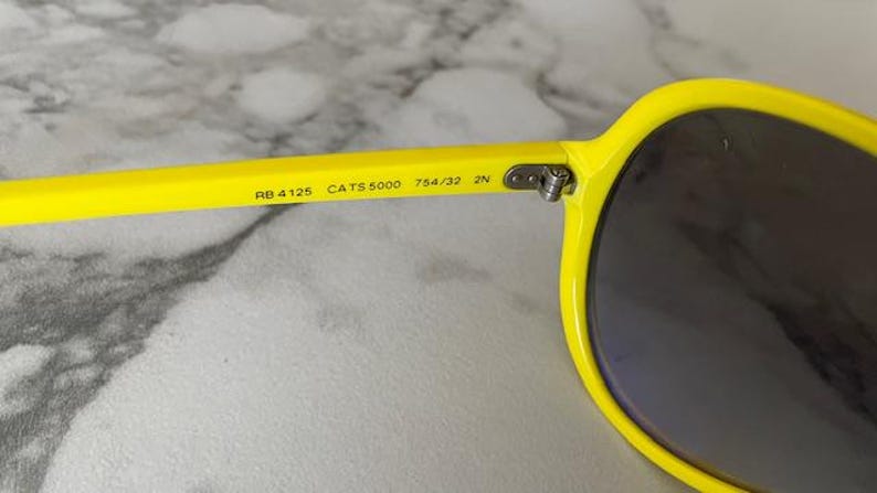May include: Close-up of bright yellow sunglasses with dark lenses. The arm of the glasses displays the text "RB 4125 CATS 5000 754/32 2N". The sunglasses have a classic, rounded shape.