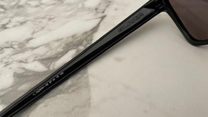 May include: Close-up of black sunglasses with a dark lens. The arm of the sunglasses has the brand name "SELIVER" printed on it. The arm also has the numbers "KL 009341-06 57 18 140" printed on it.