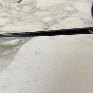 Puede incluir: Gafas de sol negras con montura gruesa y lentes oscuras. El logotipo de Ray-Ban es visible en la patilla. Las gafas de sol est&aacute;n sobre una superficie de m&aacute;rmol blanco y gris.