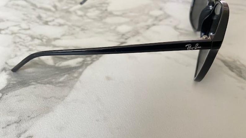 Puede incluir: Gafas de sol negras con montura gruesa y lentes oscuras. El logotipo de Ray-Ban es visible en la patilla. Las gafas de sol est&aacute;n sobre una superficie de m&aacute;rmol blanco y gris.