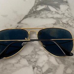 May include: Aviator-style sunglasses with a gold-colored metal frame and dark blue lenses. The sunglasses have a double bridge and black temples. The lenses are slightly reflective, and the glasses are resting on a marble surface.