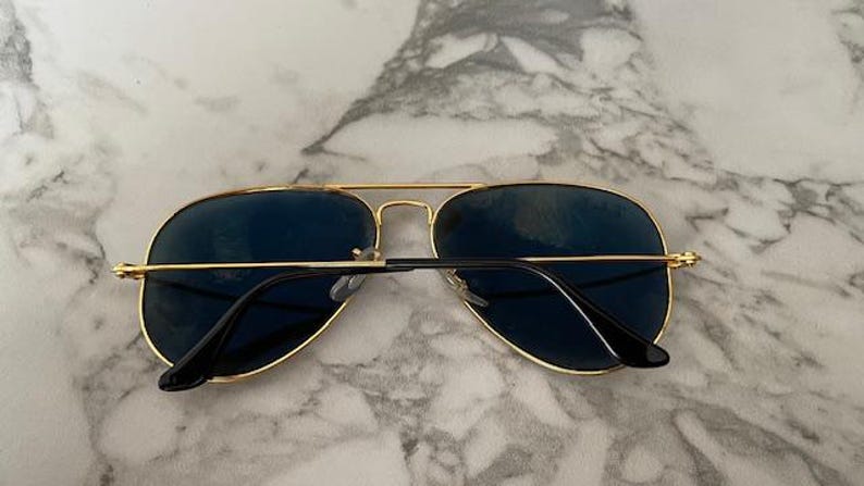 May include: Aviator-style sunglasses with a gold-colored metal frame and dark blue lenses. The sunglasses have a double bridge and black temples. The lenses are slightly reflective, and the glasses are resting on a marble surface.