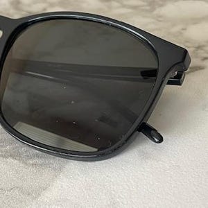 Puede incluir: Primer plano de gafas de sol negras con lentes oscuras. Las gafas tienen una montura cuadrada y est&aacute;n sobre una superficie de color claro. La imagen destaca el dise&ntilde;o elegante y la calidad reflectante de las lentes.