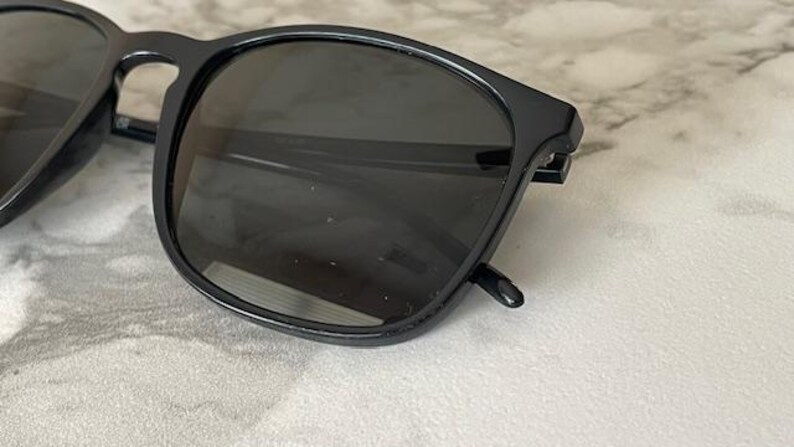 Puede incluir: Primer plano de gafas de sol negras con lentes oscuras. Las gafas tienen una montura cuadrada y est&aacute;n sobre una superficie de color claro. La imagen destaca el dise&ntilde;o elegante y la calidad reflectante de las lentes.