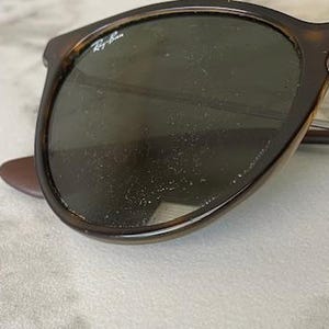 K&ouml;nnte beinhalten: Nahaufnahme einer Sonnenbrille mit Schildpattrahmen und dunklen Gl&auml;sern. Das Ray-Ban-Logo ist auf dem linken Glas zu sehen. Die Sonnenbrille liegt auf einer hellen, strukturierten Oberfl&auml;che.