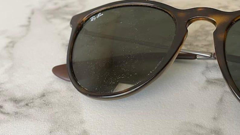 K&ouml;nnte beinhalten: Nahaufnahme einer Sonnenbrille mit Schildpattrahmen und dunklen Gl&auml;sern. Das Ray-Ban-Logo ist auf dem linken Glas zu sehen. Die Sonnenbrille liegt auf einer hellen, strukturierten Oberfl&auml;che.