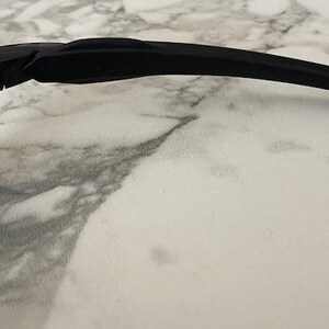 Puede incluir: Primer plano de gafas de sol negras con una patilla curva. Las lentes tienen un tinte verde. Las gafas de sol est&aacute;n sobre una superficie de m&aacute;rmol blanco y gris. La imagen est&aacute; bien iluminada.