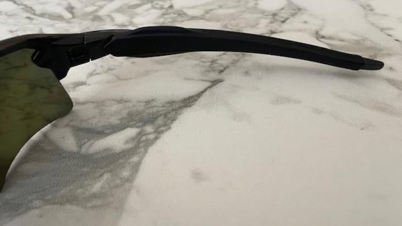 Puede incluir: Primer plano de gafas de sol negras con una patilla curva. Las lentes tienen un tinte verde. Las gafas de sol est&aacute;n sobre una superficie de m&aacute;rmol blanco y gris. La imagen est&aacute; bien iluminada.