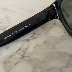 Puede incluir: Primer plano de gafas de sol negras con lentes oscuras. El texto "RB 4194 601/BA 53o17 140 3P" est&aacute; impreso en la patilla de las gafas de sol. Las gafas de sol descansan sobre una superficie clara y veteada.
