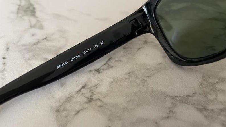 Puede incluir: Primer plano de gafas de sol negras con lentes oscuras. El texto "RB 4194 601/BA 53o17 140 3P" est&aacute; impreso en la patilla de las gafas de sol. Las gafas de sol descansan sobre una superficie clara y veteada.