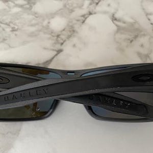 Może przedstawiać: Czarne okulary przeciwsłoneczne marki Oakley z prostokątną oprawką i ciemnymi soczewkami. Nazwa marki jest nadrukowana na zausznikach. Okulary są złożone.