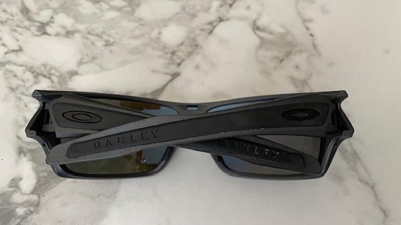 Może przedstawiać: Czarne okulary przeciwsłoneczne marki Oakley z prostokątną oprawką i ciemnymi soczewkami. Nazwa marki jest nadrukowana na zausznikach. Okulary są złożone.