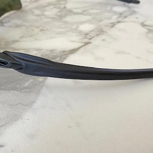 Puede incluir: Gafas de sol deportivas negras y grises con un dise&ntilde;o curvo. Las gafas tienen una montura y lentes oscuras, con un logotipo plateado en el lateral. Las gafas est&aacute;n sobre un fondo de m&aacute;rmol blanco y gris.