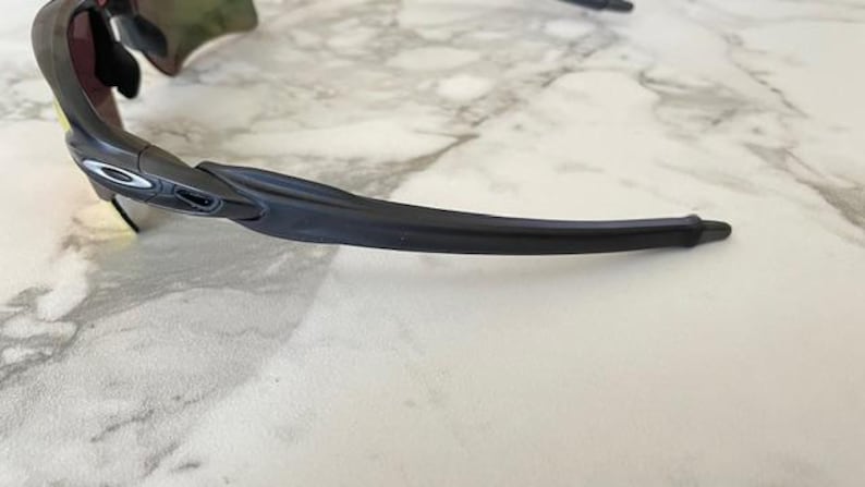 Puede incluir: Gafas de sol deportivas negras y grises con un dise&ntilde;o curvo. Las gafas tienen una montura y lentes oscuras, con un logotipo plateado en el lateral. Las gafas est&aacute;n sobre un fondo de m&aacute;rmol blanco y gris.